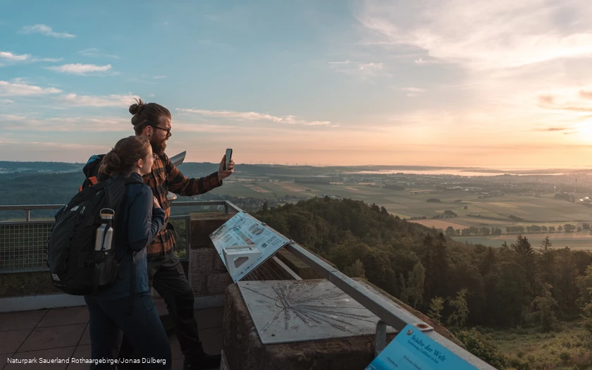Ein Wanderpaar steht bei Sonnenaufgang auf dem Georg-Victor Turm mit dem Handy in der Hand