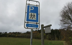 Parkplatzschild mit weißem P auf blauem Grund und darunter zwei Männchen mit einem Wanderstock und der Aufschrift Am Rees.