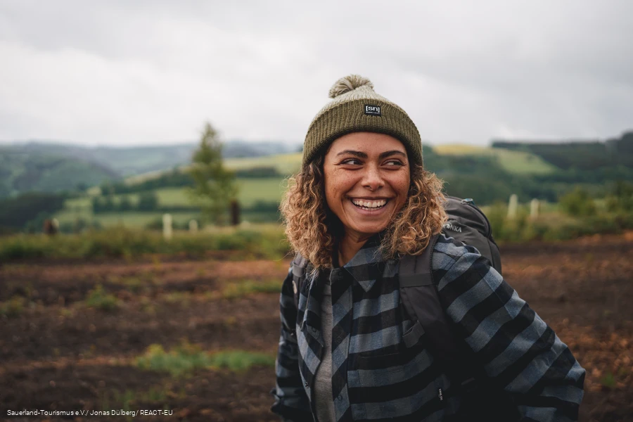 Eine Frau steht in Wanderausr&uuml;stung in einer idyllischen Landschaft und ist sichtlich gl&uuml;cklich